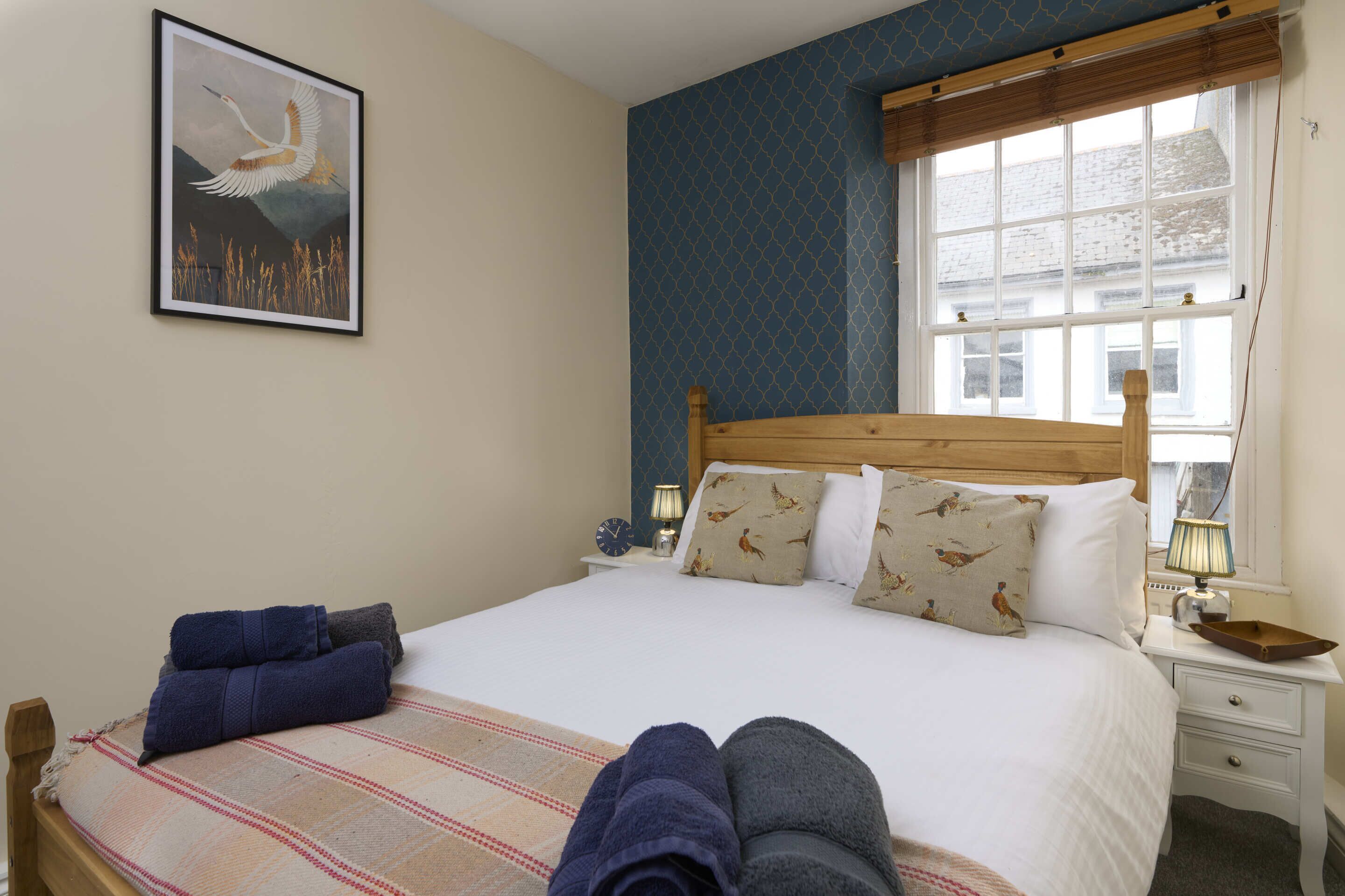 A neatly made bed with a plaid bedspread, a framed picture of a bird on the wall, and a window with a view of buildings.