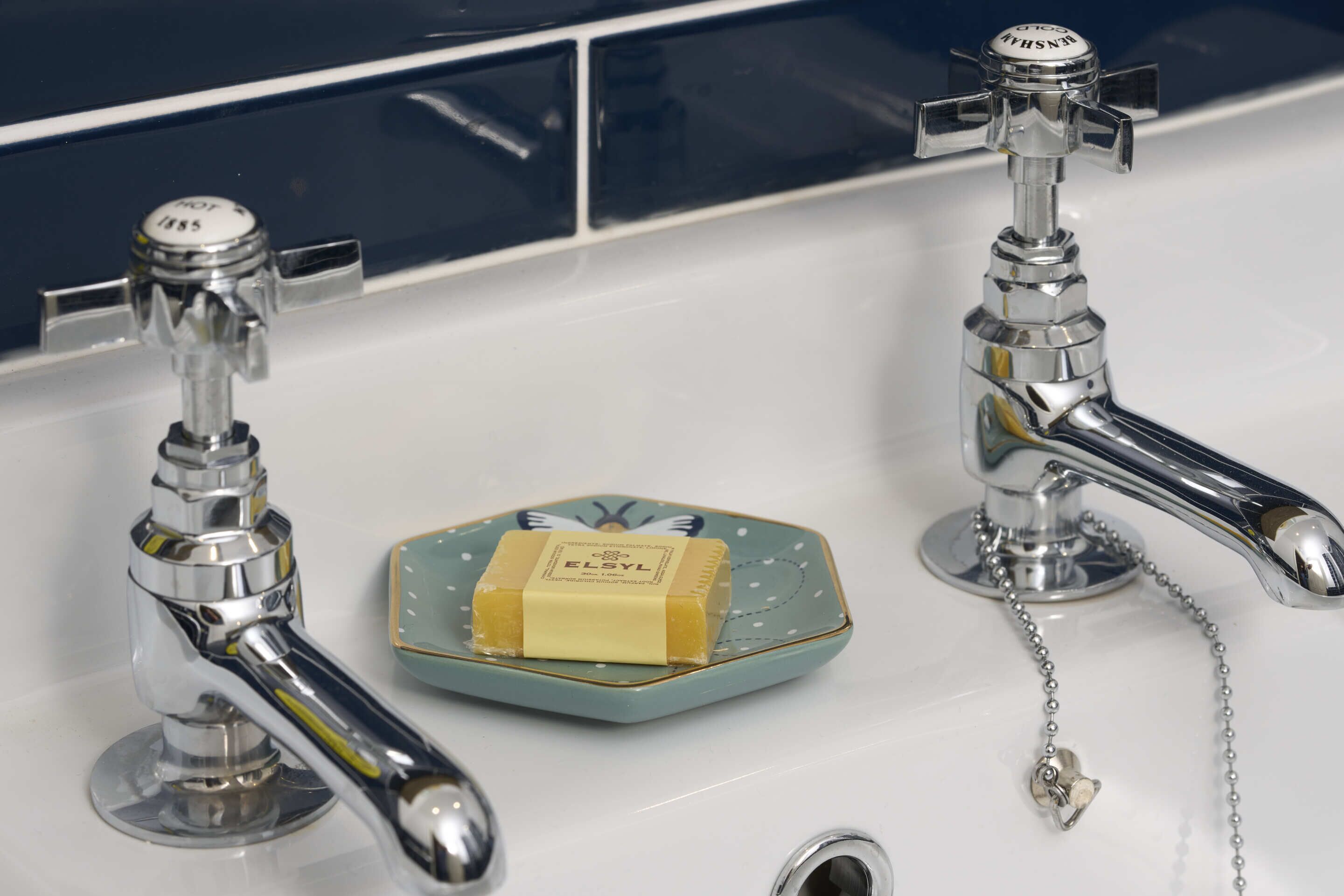 Two chrome faucets on a white sink with a soap bar on a decorative dish.