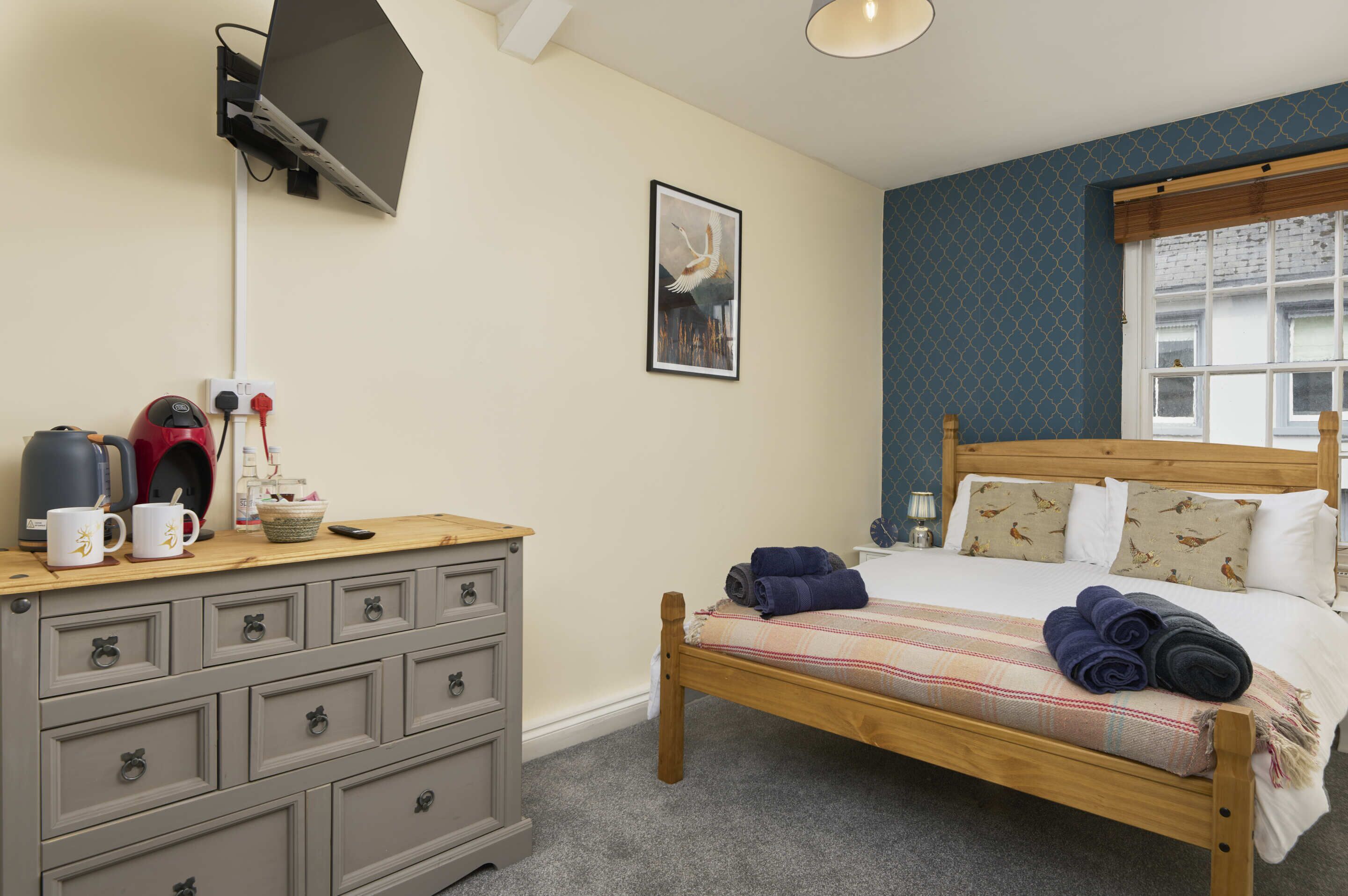 Een slaapkamer met een houten bed, een dressoir met lades, een flatscreen televisie aan de muur en een raam met gordijnen met patroon.