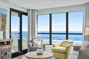 A room with a large window overlooking the ocean, a round wooden table with a decorative item, and two armchairs.
