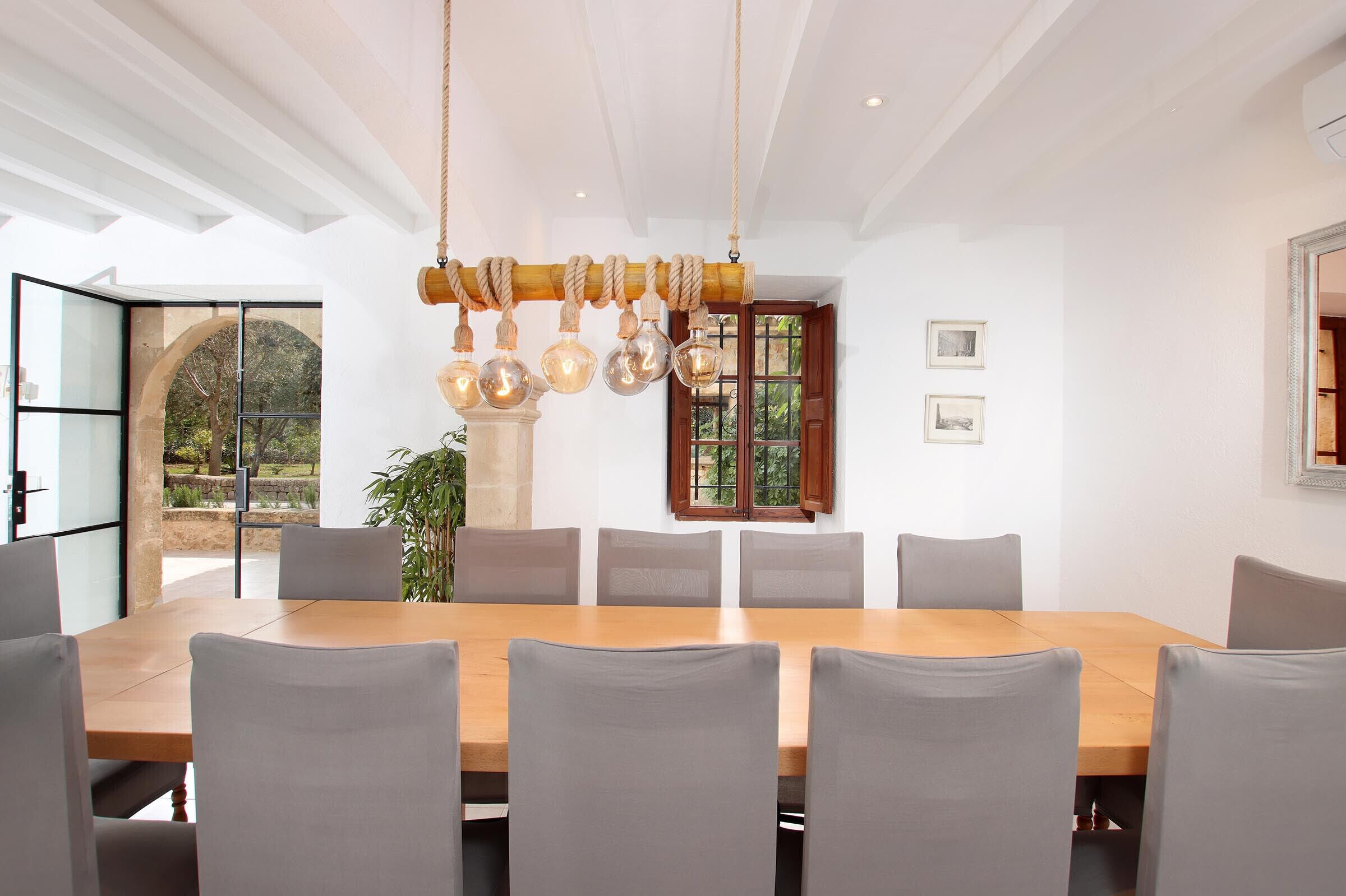 A dining area with a wooden table and grey chairs, a hanging light fixture, and a window with a view of greenery.