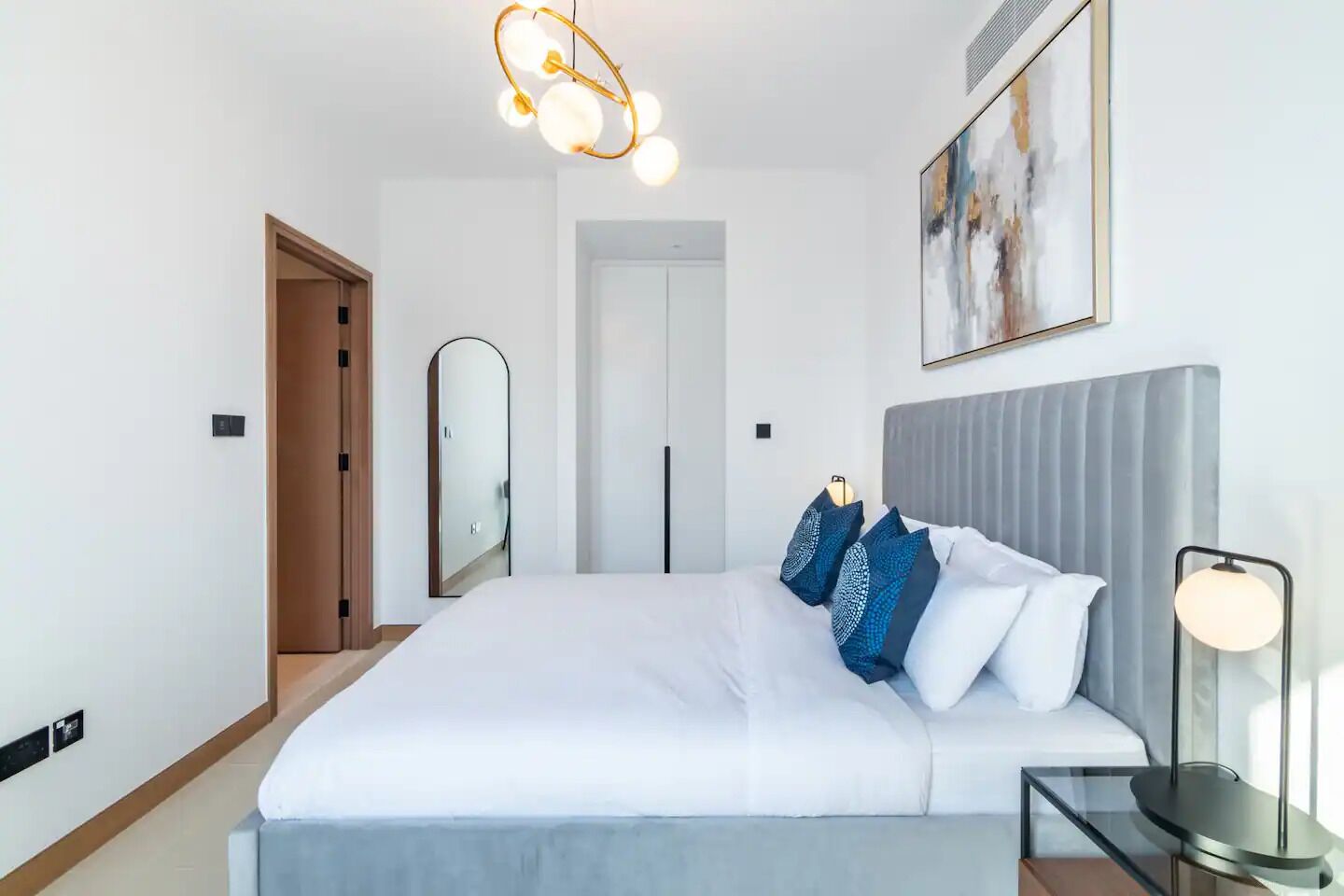 A modern hotel room with a large bed, a grey headboard, a bedside table with a lamp, and a painting on the wall.