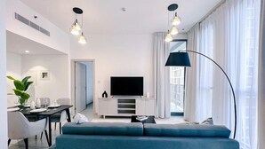 A modern living room with a large blue sofa, a flat-screen TV, and a dining area with a table and chairs. - Green community 1BR 15 min to sea (Dubai)