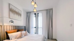 A modern hotel room with a large bed, a headboard, a bedside lamp, and a view of the outdoors through sheer curtains. - Green community 1BR 15 min to sea (Dubai)