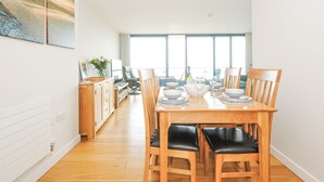 Un comedor con una mesa de madera dispuesta para cuatro, flanqueada por dos sillas de madera con cojines negros. Se ve una cocina al fondo, con una silla gris y un florero sobre la encimera.