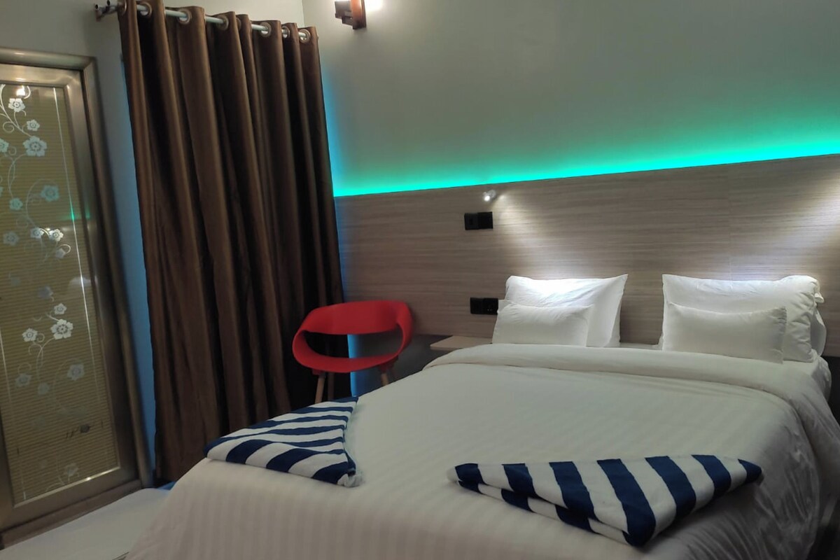 A hotel room with a bed, a nightstand, a red chair, and a wall with a light strip.
