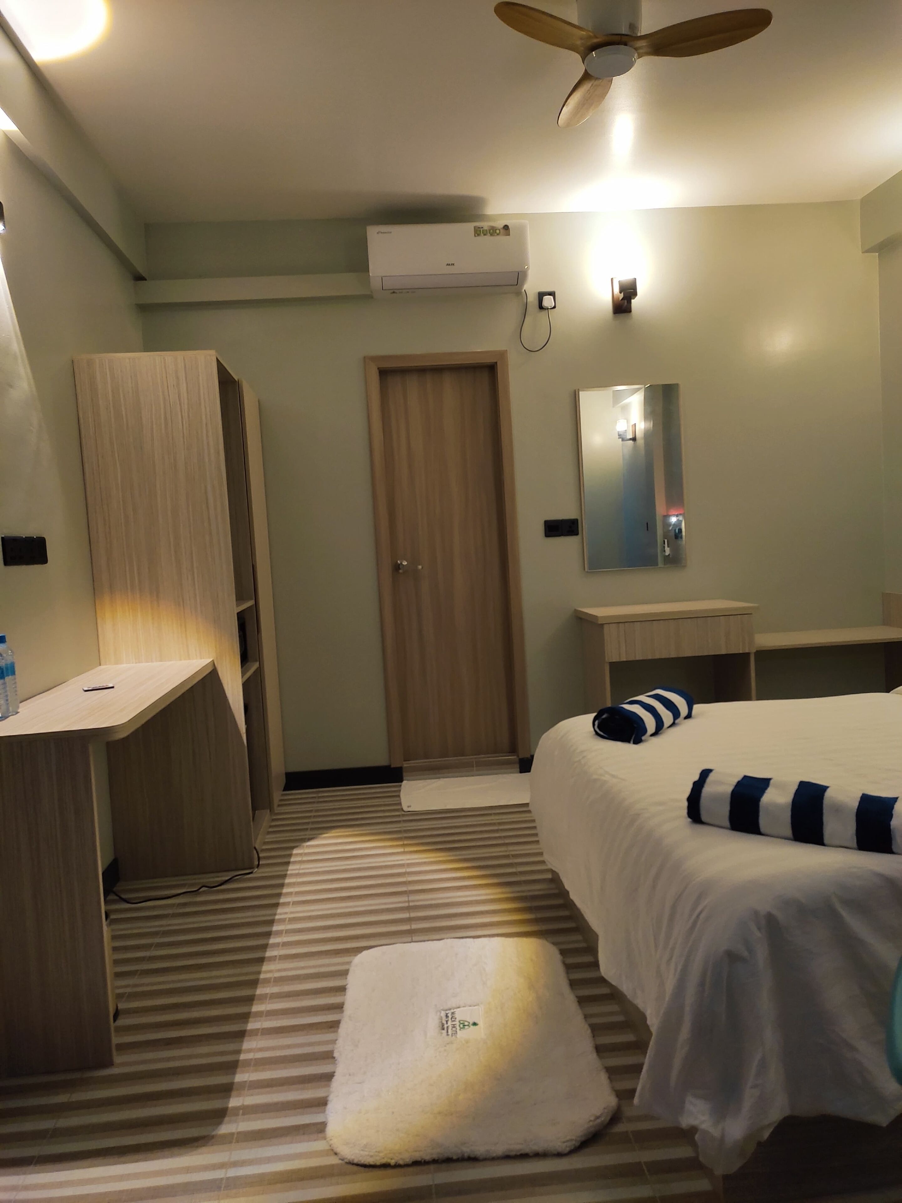 A hotel room with a bed, a desk, a wardrobe, a mirror, and a ceiling fan.