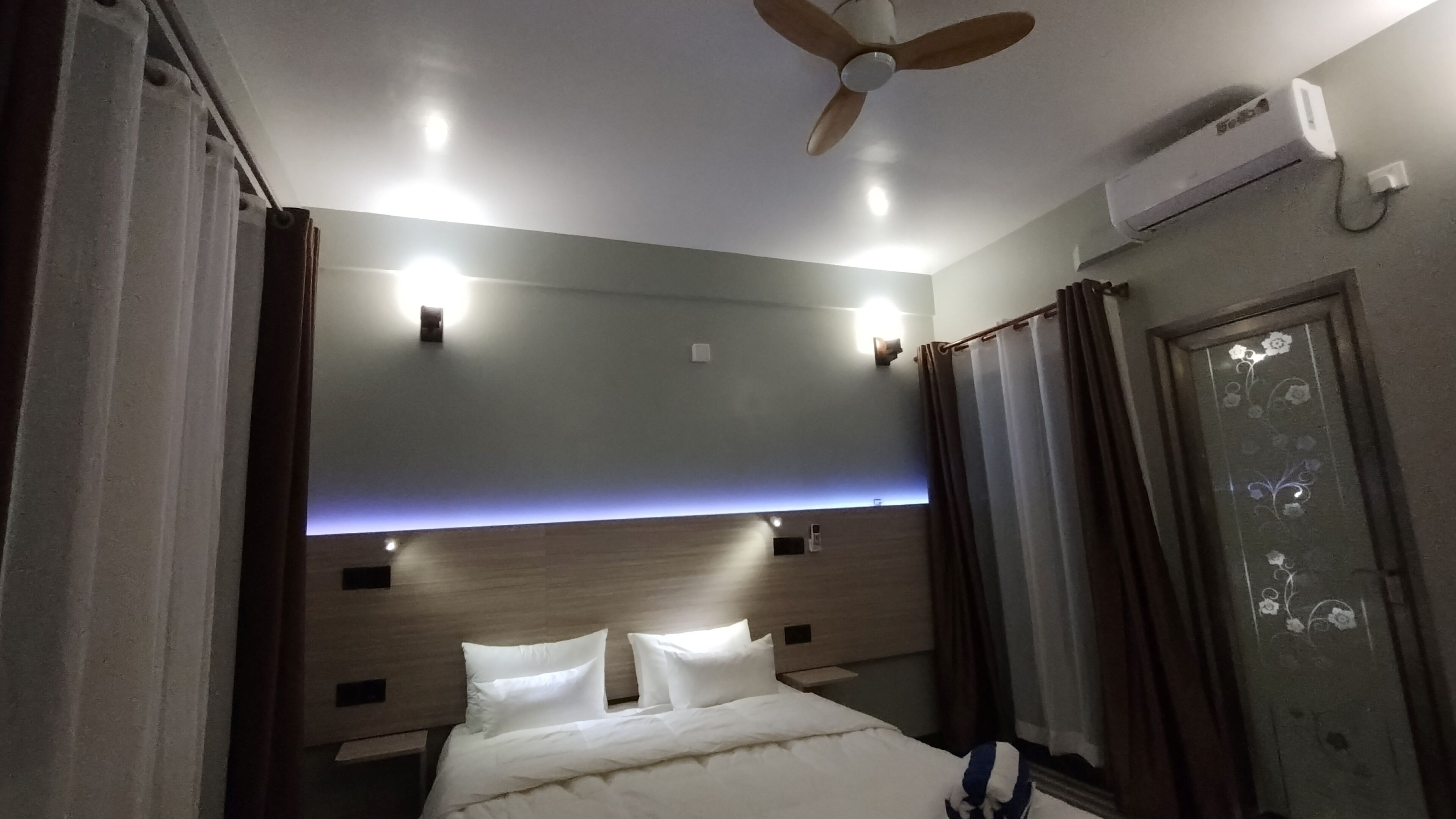 A hotel room with a bed, ceiling fan, air conditioning unit, and a nightstand.