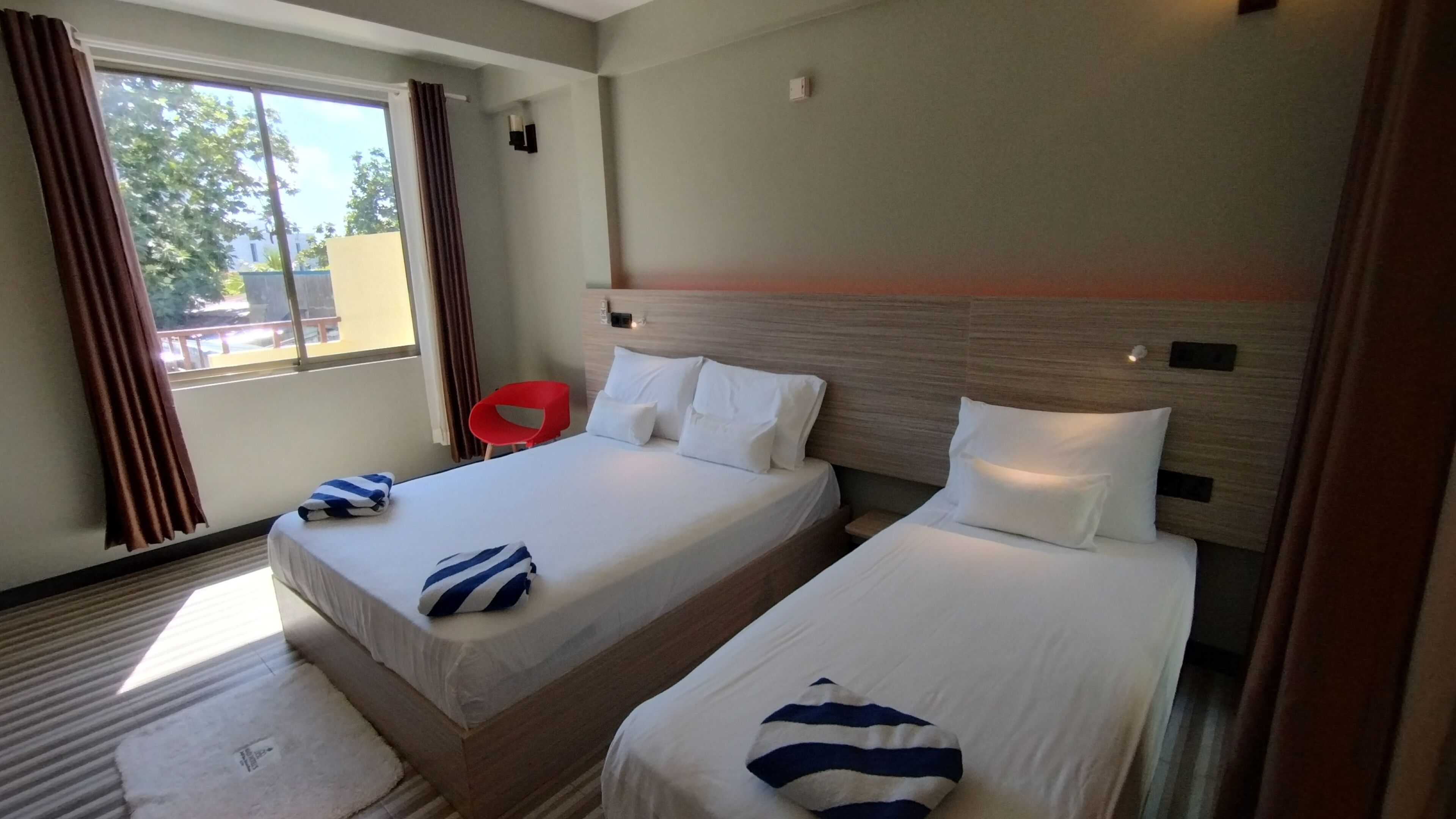 A hotel room with two beds, a red chair, and a view of the outside.