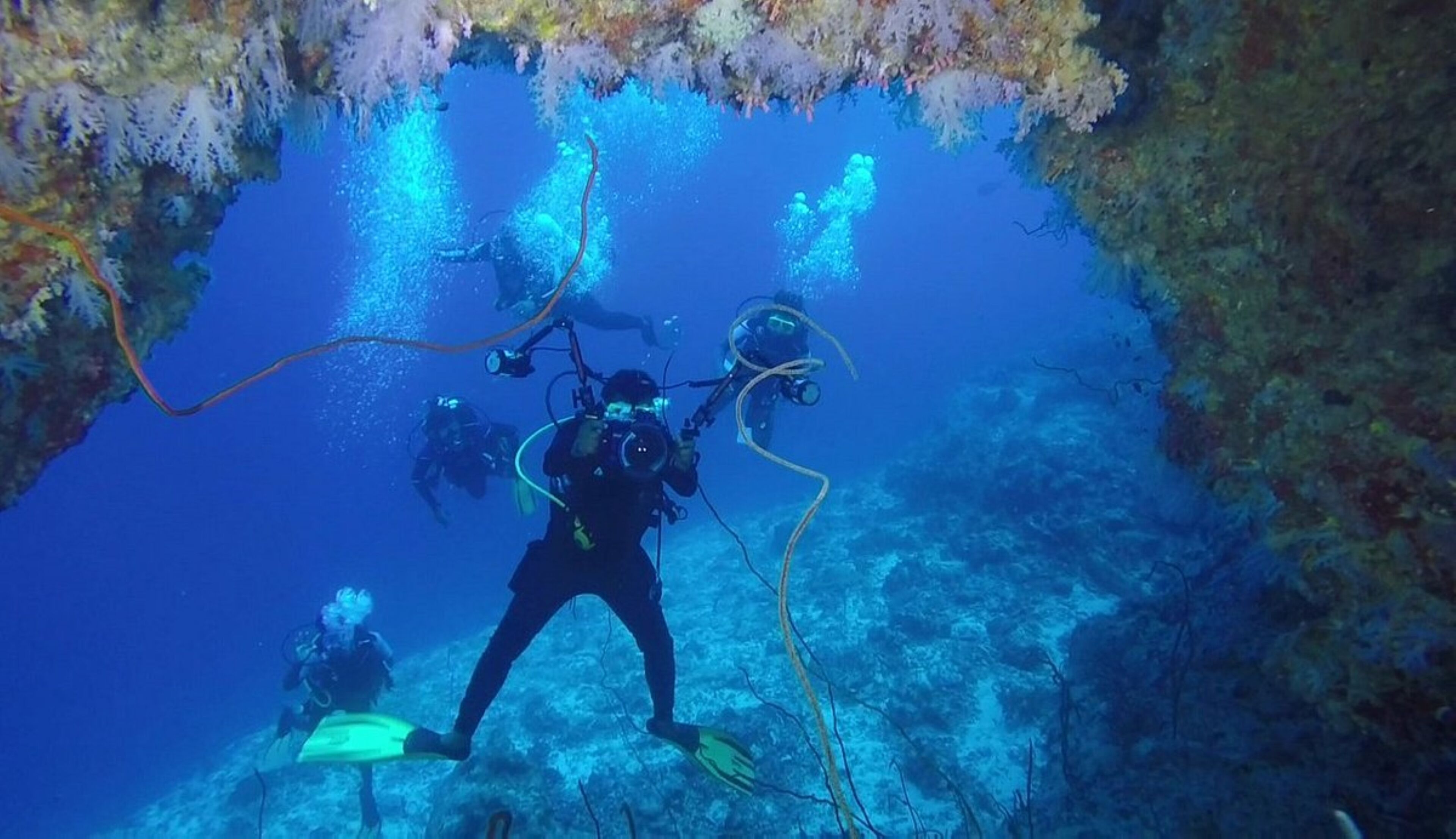 Divers exploring an underwater cave with visible coral formations and rocky terrain.