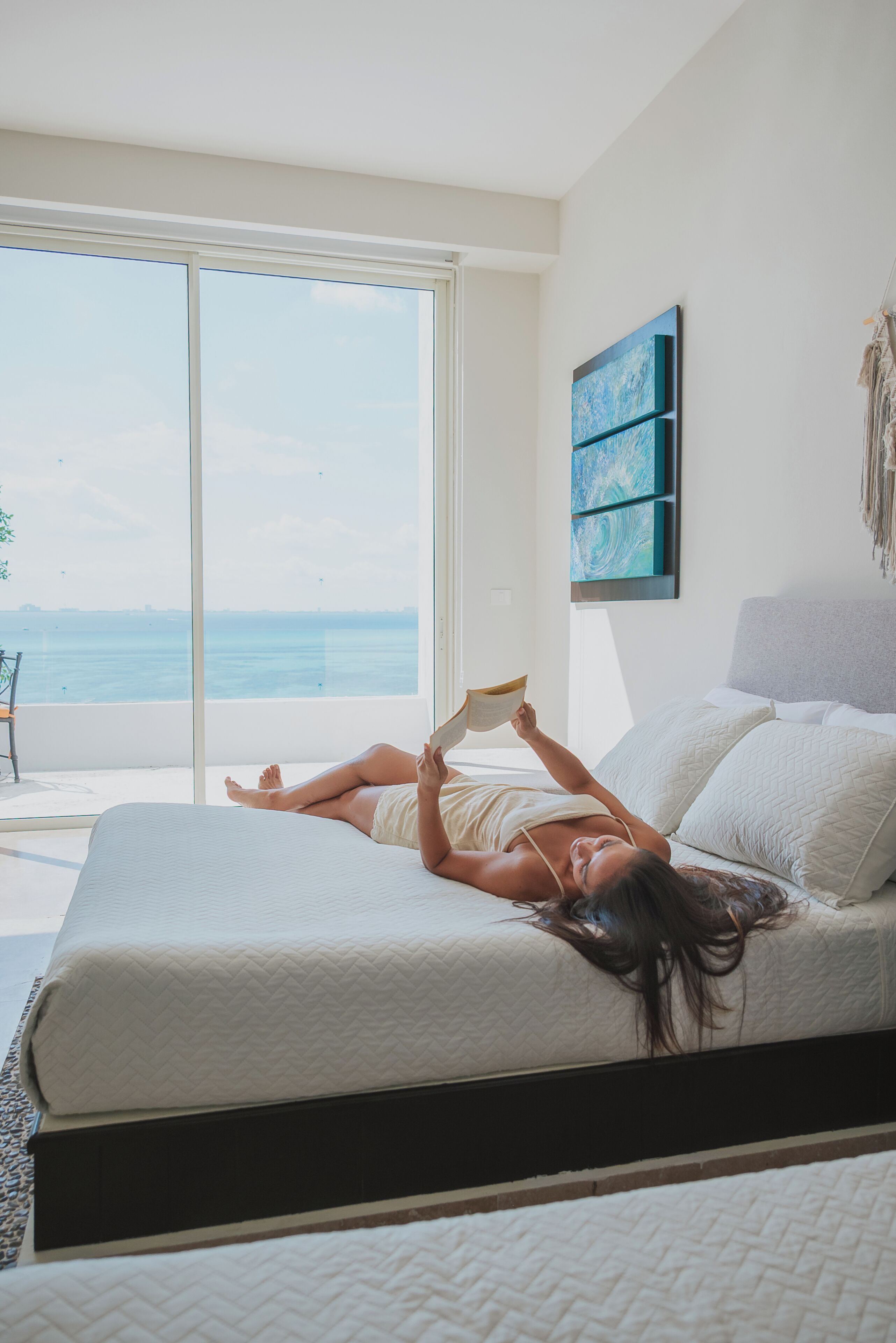 Een slaapkamer met een groot raam dat uitkijkt op de zee, een bed en een kunstwerk aan de muur.
