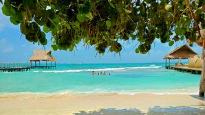 A beach with clear turquoise water, a wooden pier, and thatched-roof huts. - Villa Chan Kah (Isla Mujeres)