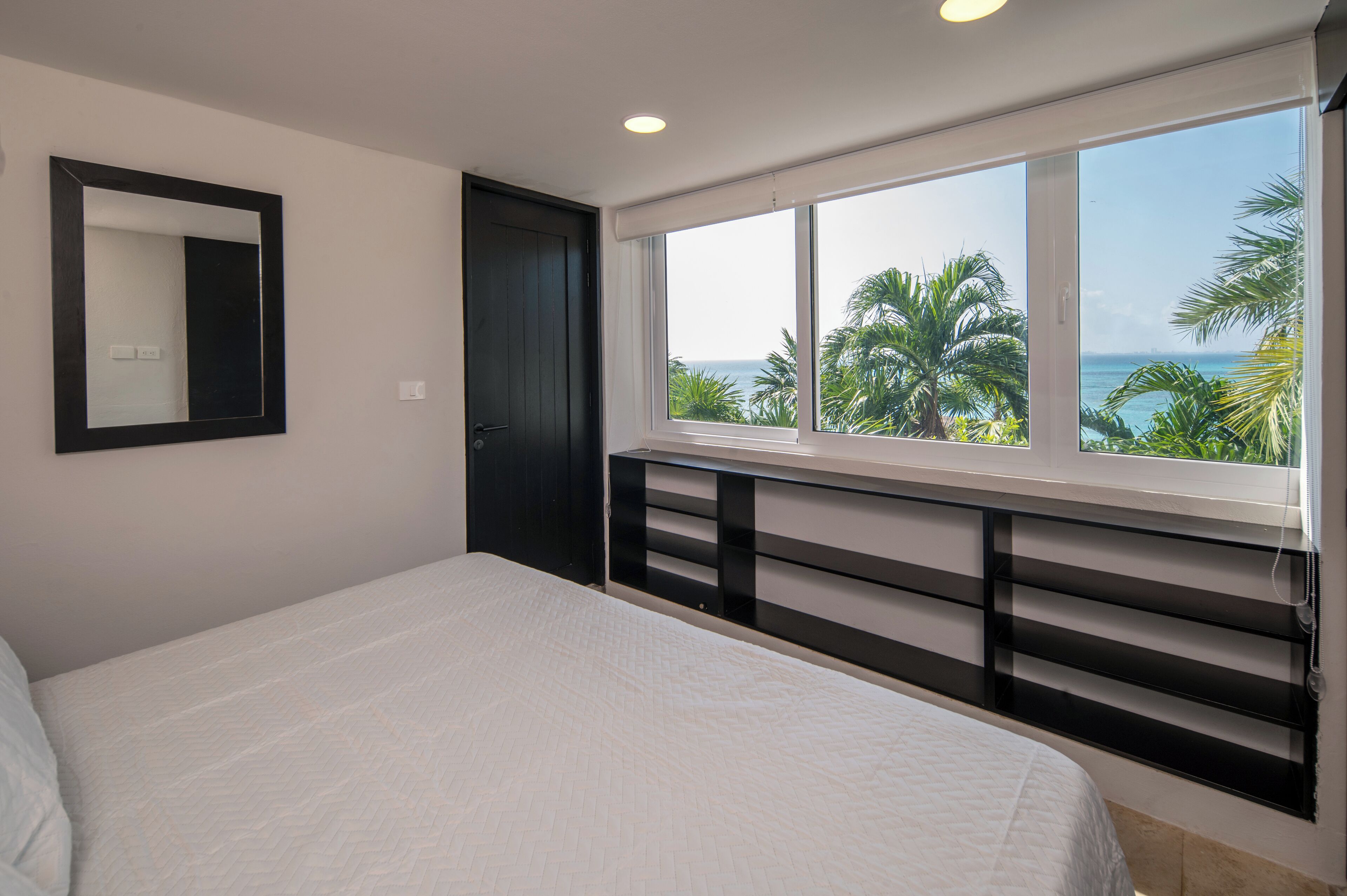 Une chambre avec un grand lit, une fenêtre donnant sur des palmiers et la mer, une porte sombre et un miroir sur le mur.