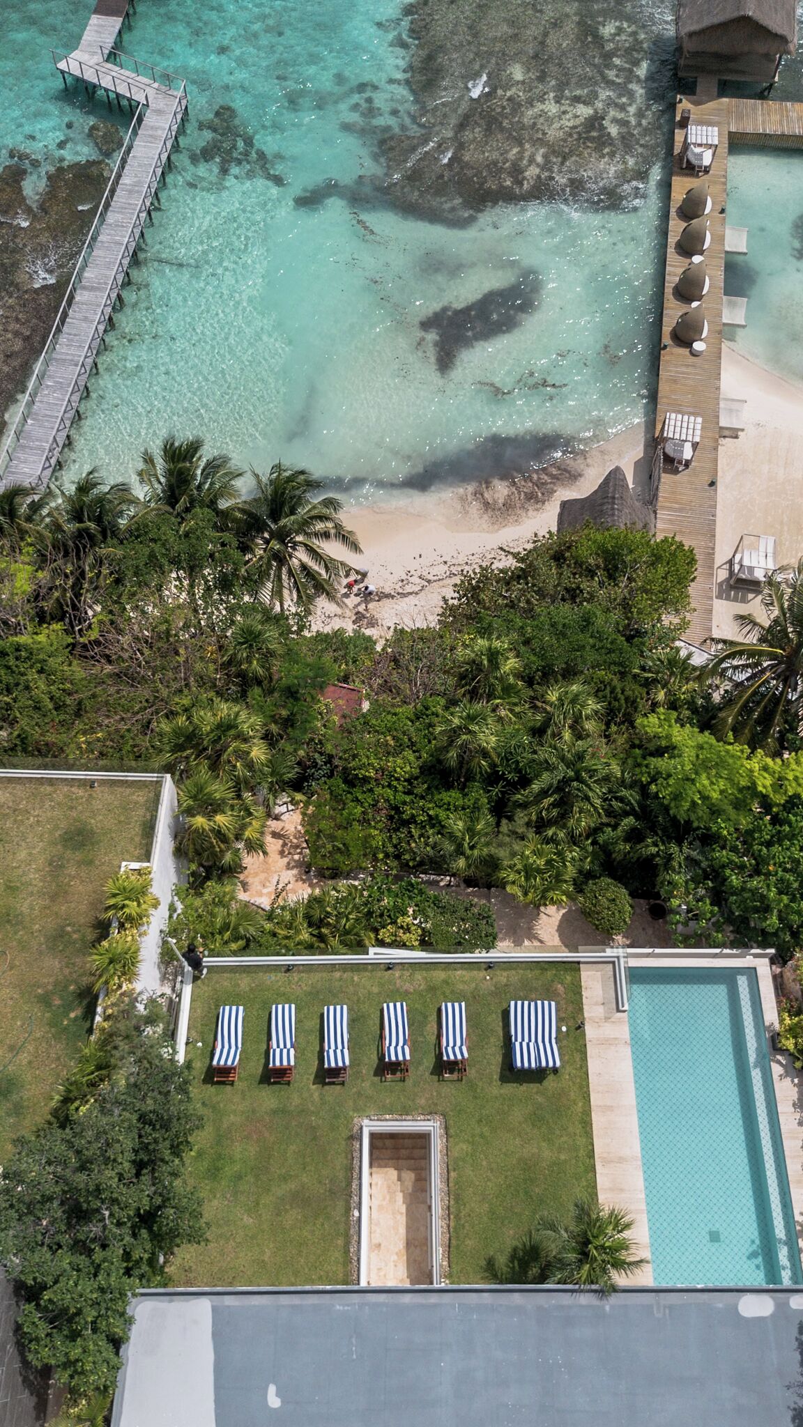 Ein Luftbild eines Resorts mit Strand, einem hölzernen Steg und einem Poolbereich mit Liegestühlen.