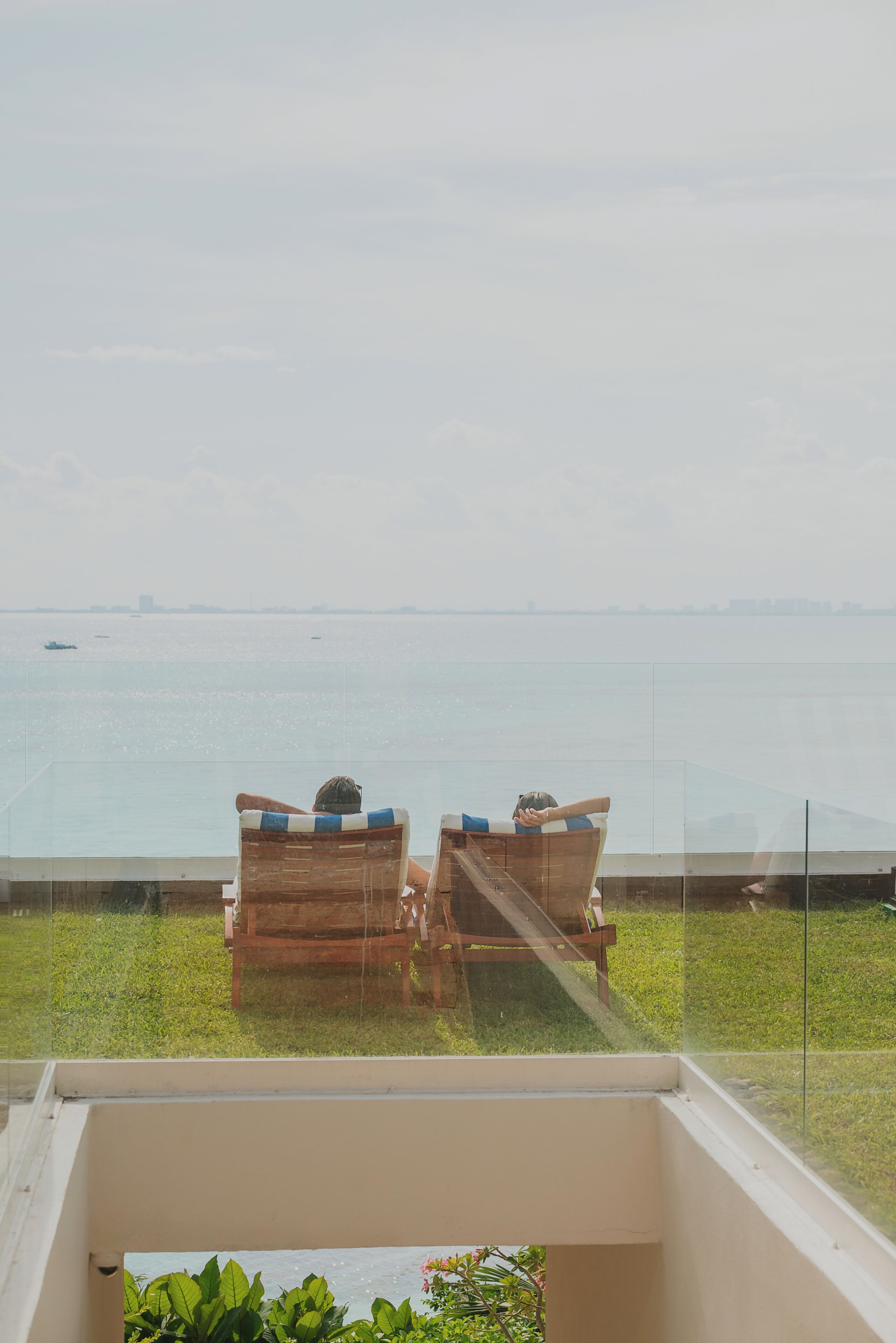 Dos sillas de madera mirando un balcón de barandilla de cristal con vista a un cuerpo de agua y barcos a lo lejos.