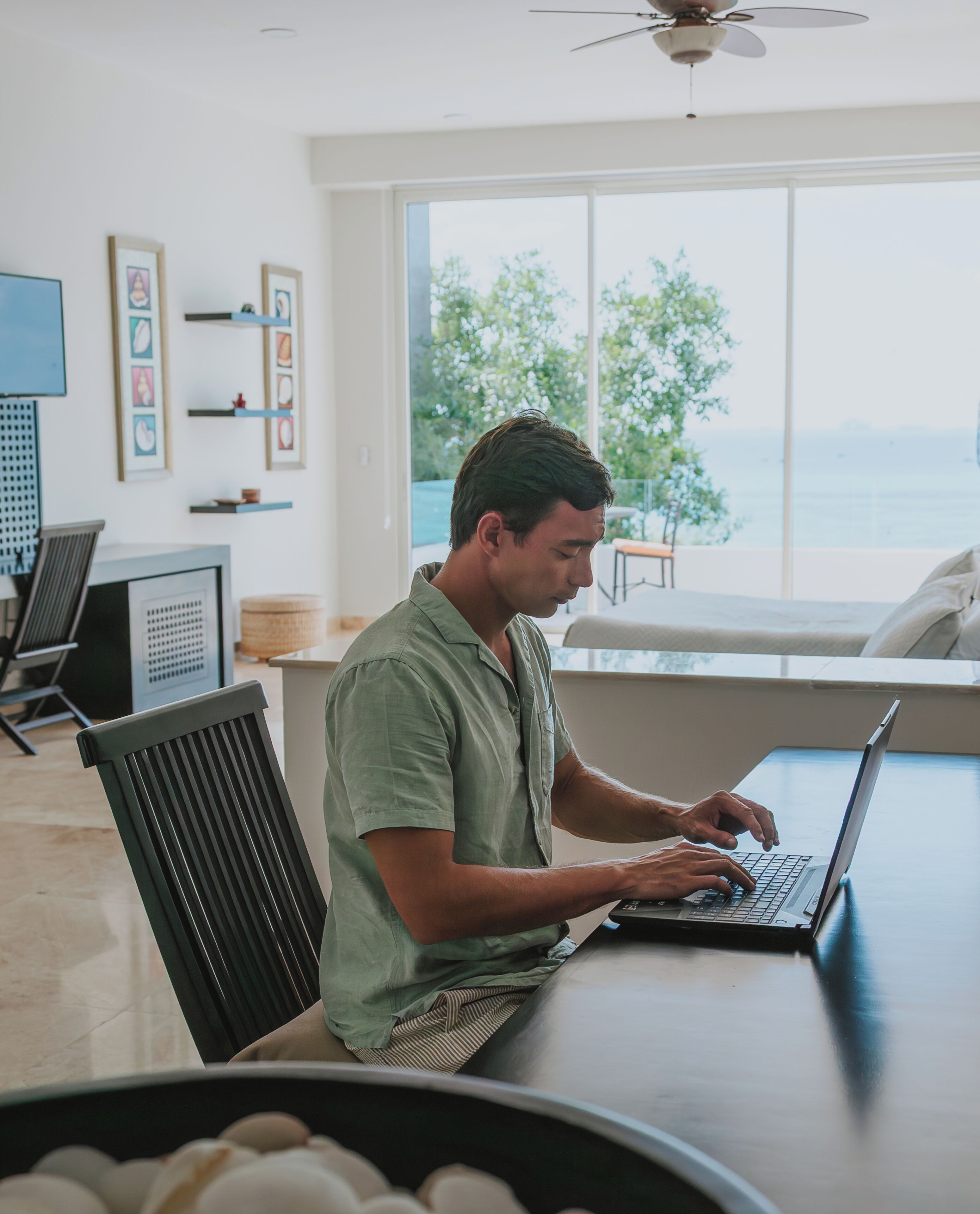 A person working on a laptop in a room with a large window, a ceiling fan, and framed pictures on the wall.