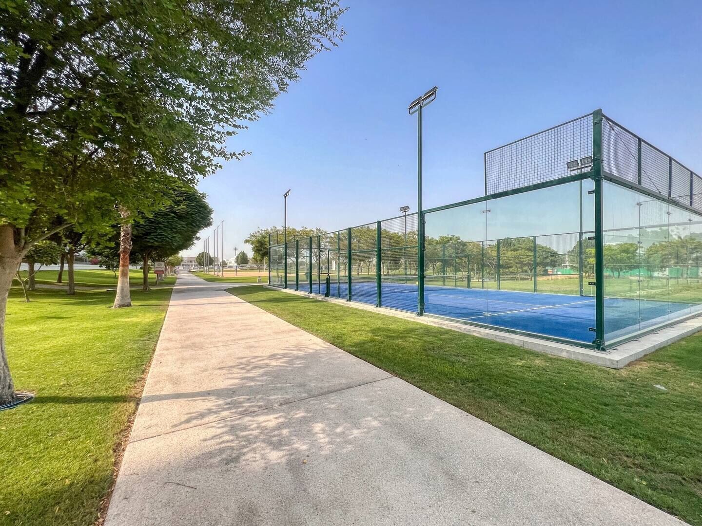 Un court de tennis entouré d’une paroi en verre, bordé d’une allée en béton et d’arbres.