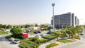 A modern urban area with a multi-story building, a parking lot, and a street with cars and a red container labeled "MYBIKE". - Cozy studio at Damac Hills (Dubai)