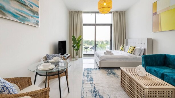 A modern hotel room with a bed, a blue sofa, a glass dining table, and a large window. - Cozy studio at Damac Hills (Dubai)
