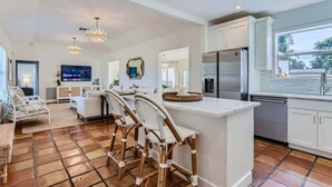 A modern kitchen with white cabinetry, a central island with bar stools, a large window, and a flat-screen TV mounted on the wall. - The Glenn House - Gather Poolside in a Breezy South End Getaway (West Palm Beach)