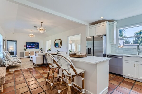 A modern kitchen with white cabinetry, a central island with bar stools, a built-in refrigerator, and a large window providing a view of the outdoors.