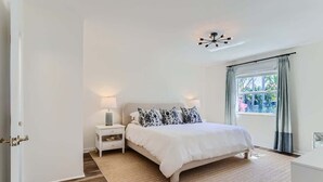 A bedroom with a bed, nightstands, a lamp, and a window with curtains. - The Glenn House - Gather Poolside in a Breezy South End Getaway (West Palm Beach)