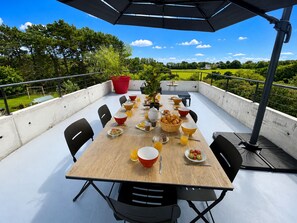 An outdoor dining area with a table set for breakfast, surrounded by chairs, and an umbrella providing shade. - La Villa Océane, Elegante, Plages, Gr34, 2-8 Pers (Clohars-Carnoët)