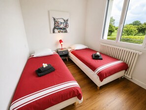 A hotel room with two beds, a nightstand, a lamp, a painting, and a window with a view of greenery. - La Villa Océane, Elegante, Plages, Gr34, 2-8 Pers (Clohars-Carnoët)