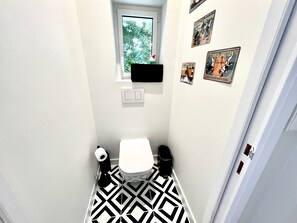 Un bagno stretto con pavimento a motivi bianco e neri, un water, una piccola finestra e diverse immagini appese alle pareti.