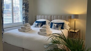 Ein ordentlich bezogenes Bett mit weißen Laken und blauen Kissen, eine Nachttischlampe und ein Fenster mit Vorhängen.