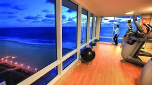 A gym with exercise equipment and a large window overlooking a beach at dusk. - Oceanfront Luxury at Hilton Oceanaire Resort – Two Stunning Oceanview Balconies! (Virginia Beach)