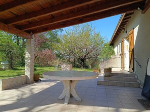 A covered patio area with a round table, a stone bench, and a view of a building with wooden doors and a stone wall. - Charming family home in Réalmont - Near Albi (UNESCO) (Réalmont)