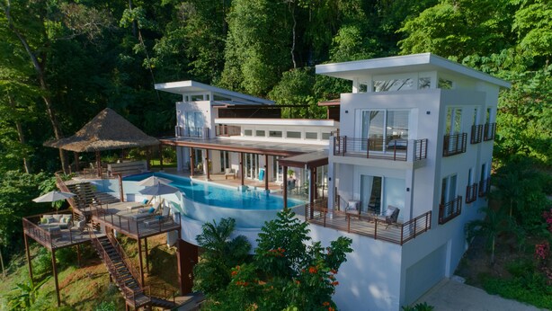 A modern multi-story house with a swimming pool, outdoor seating, and a thatched-roof structure.