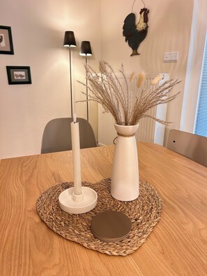 A wooden dining table with a woven placemat, a white vase with dried plants, and two floor lamps. - House Meerten (Neroth)