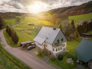 A two-story house with a sloped roof, a car parked in front, and a small garden with a flower bed. - House Meerten (Neroth)