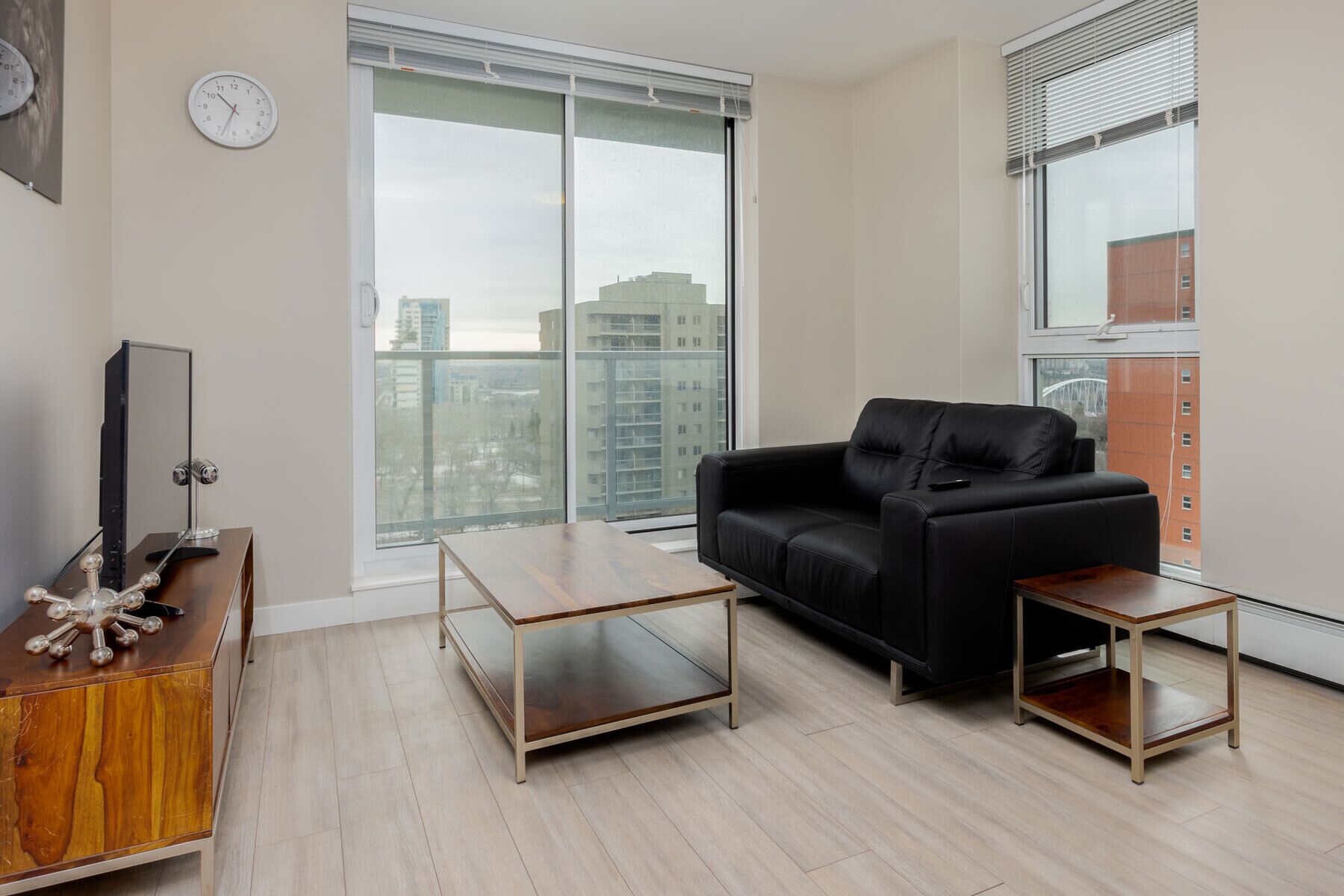 Een moderne woonkamer met een zwarte leren bank, een houten salontafel en een bijzettafeltje. Er is een groot raam met uitzicht op gebouwen en een stadsgezicht.