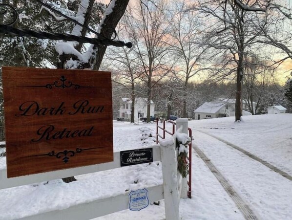 Un panneau en bois indiquant « Dark Run Retreat », avec un panneau plus petit en dessous portant la mention « Résidence privée », le tout placé sur un fond de paysage enneigé, avec des arbres sans feuilles et une maison au loin.