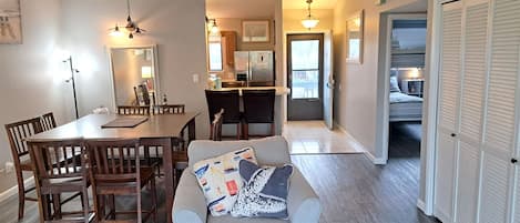 A dining area with a wooden table and chairs, a gray sofa with patterned cushions, and a kitchen in the background.