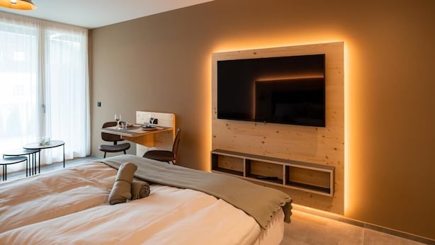 A modern hotel room with a large bed, a desk with a chair, a flat-screen TV mounted on a wooden wall, and a small table with a chair. - Holiday apartment Ammerglück "Classic" in Oberammergau (Oberammergau)