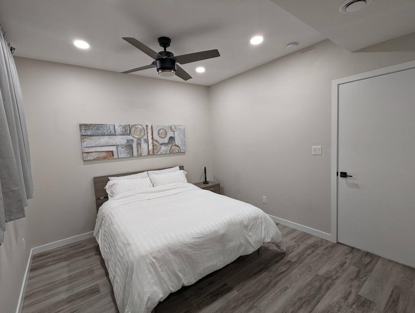 Ein Schlafzimmer mit einem Bett, einem Deckenventilator, Einbauleuchten, einer Tür und einem Wandbild.