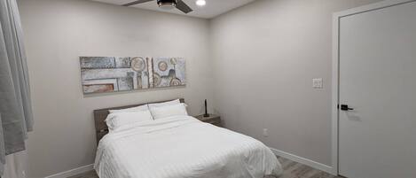 Ein Schlafzimmer mit einem Bett, einem Deckenventilator, Einbauleuchten, einer Tür und einem Wandbild.
