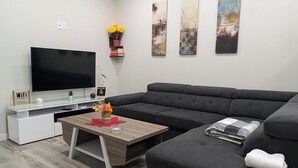A modern living room with a flat-screen TV, a dark grey sofa, a wooden coffee table, and a small side table with a vase of flowers. - Confortable brand-new 1-bedroom close to downtown
(Saskatoon)