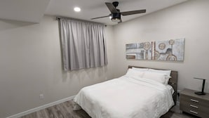 A bedroom with a bed, a ceiling fan, a window with curtains, a bedside table, and a wall-mounted artwork. - Confortable brand-new 1-bedroom close to downtown
(Saskatoon)