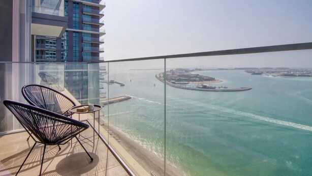A balcony with two chairs and a small table, overlooking a body of water with islands and buildings in the distance. - Sea view 2BR Beach Isle (Dubai)