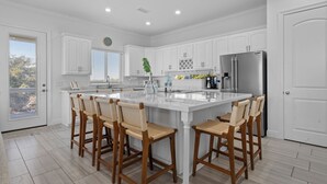 Une cuisine moderne avec des meubles blancs, un îlot central doté de tabourets, un réfrigérateur en acier inoxydable et une vue par la fenêtre.