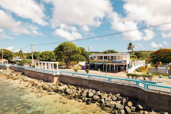 A coastal hotel with a white facade, a covered outdoor seating area, and a view of the sea.