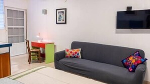 A living room with a grey sofa, colorful pillows, a small desk with a chair, and a TV mounted on the wall. - Studio #3 Anacaona w/ Garden- Guayacan Guesthouse (Vieques)