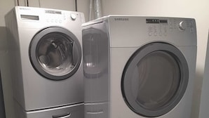 Two stacked Samsung front-loading washing machines in a room with a concrete floor and a vent pipe on the wall.