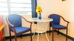 Une petite pièce avec deux fauteuils bleus, une table ronde sur laquelle est posé un vase bleu, et une fenêtre avec des rideaux.