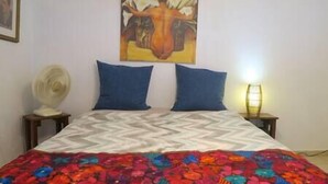 A bed with a patterned bedspread, two pillows, a wall-mounted painting, a bedside lamp, and a fan on a nightstand. - Oceanview Apartment & Garden (Vieques)